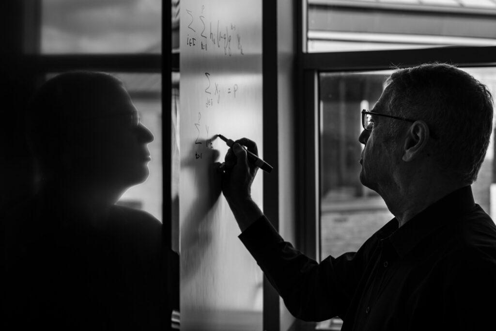 Mark Daskin writes mathematical equations on a whiteboard in his office. He is mirrored by a reflection in a window next to the whiteboard.