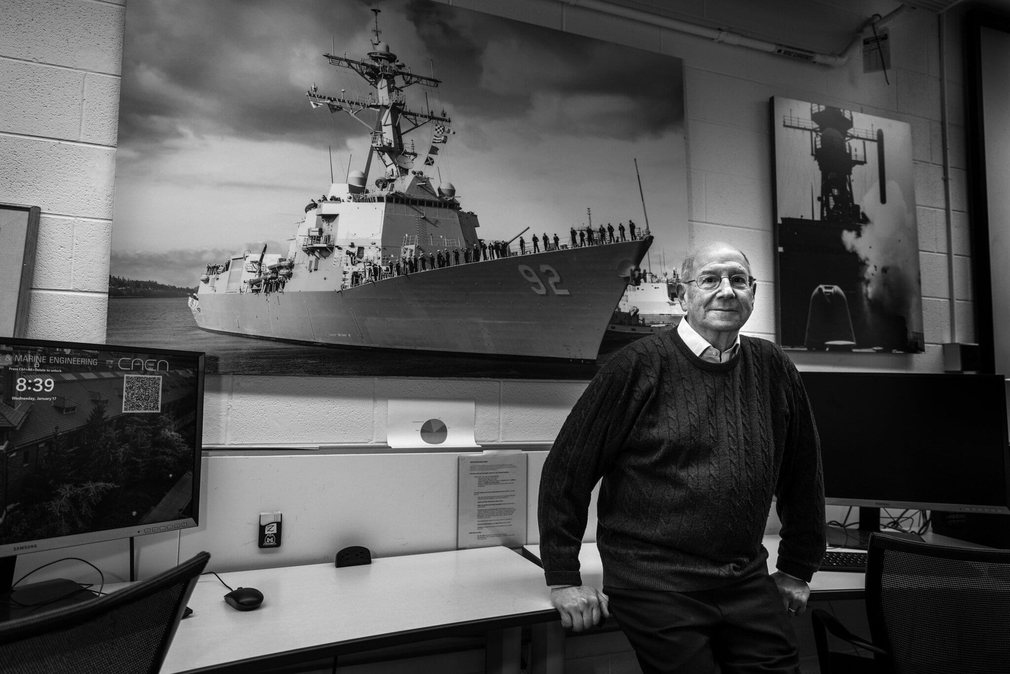 Donald Winter leans against a desk. Behind him are large photographs of naval battleships. Winter stands smiling in a sweater.