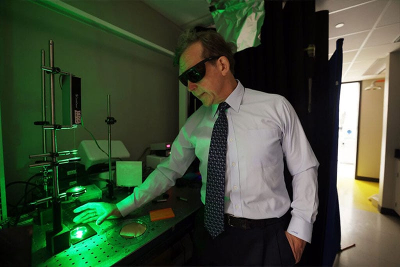 Nick Kotov wears safety glasses as he adjusts his plastic substrate. A green light illuminates the substrate in an otherwise dark room