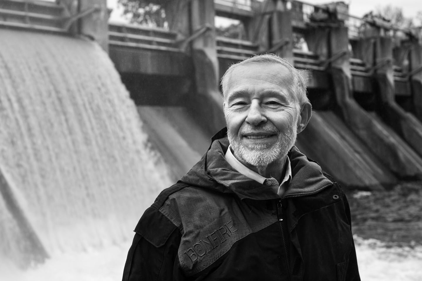 Daigger stands next to a small dam. To his back, water rushes into a body of water. Daigger is standing in a windbreaker jacket and is smiling. The image has a moody black and white treatment.