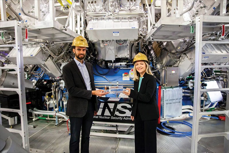 Two people stand in a metallic lab holding a target assembly. They both are wearing safety helmets and business suits.