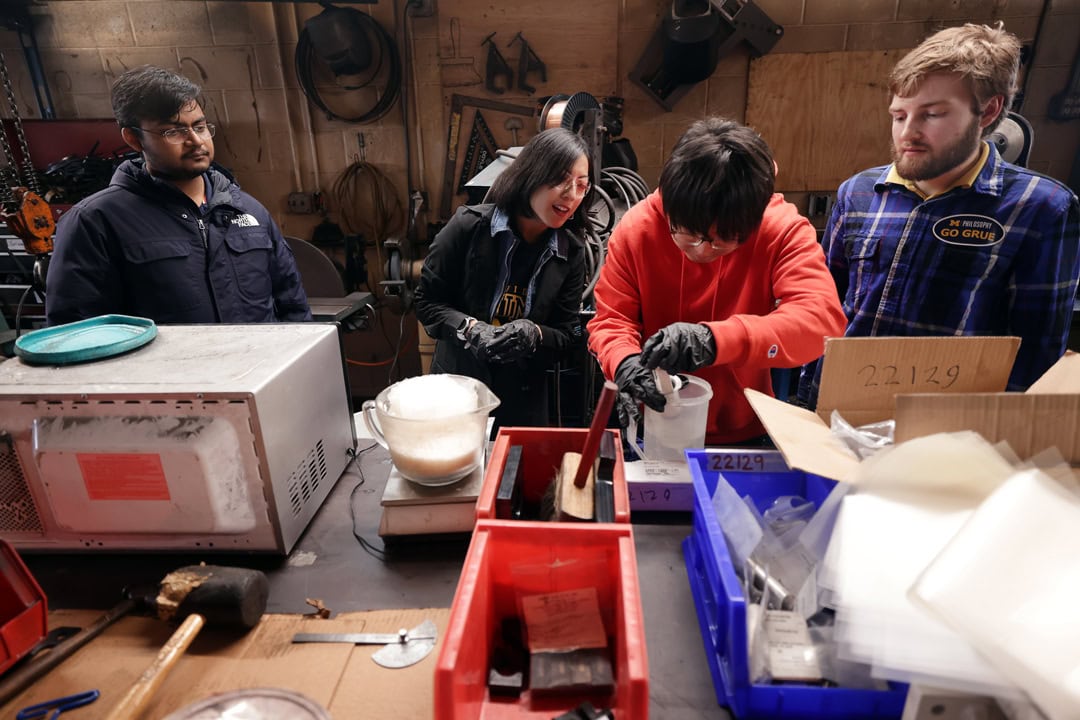 Student Jooyoung Chung measures petroleum jelly, wearing black latex gloves, onto a scale while the other students observe, inside  the Warmilu Pack Production Facility. 