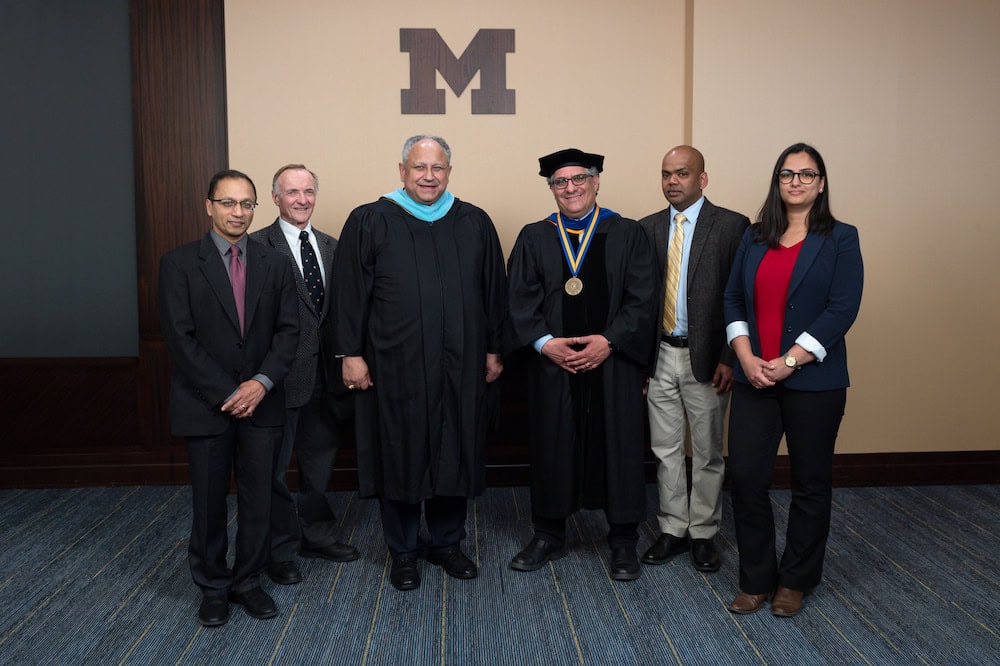 Six people stand in a line beneath the University of Michigan block M, which is printed on a wall in brown. Two men in the center of the lineup are wearing graduation regalia.