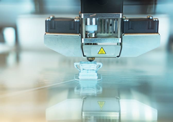 A metallic, trapezoidal-shaped 3D printer head hovers over a square, plastic object with handles on either side. A metal point protrudes from the printer head and touches the top of the plastic piece below.