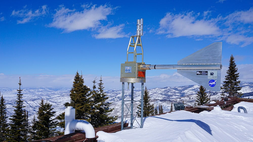 A finned cylinder protrudes from a metal box situated on a tower on the snow-capped roof of a laboratory. The lab and tower overlook a mountainous landscape covered in bright white snow.