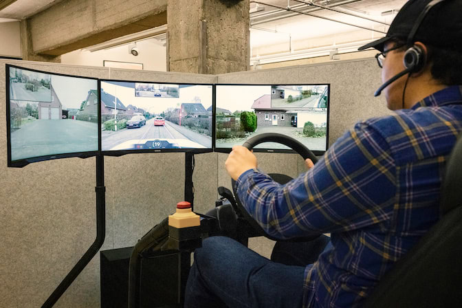 A man sits in front of a bank of three computer screens, holding a steering wheel as he pilots a remote vehicle.