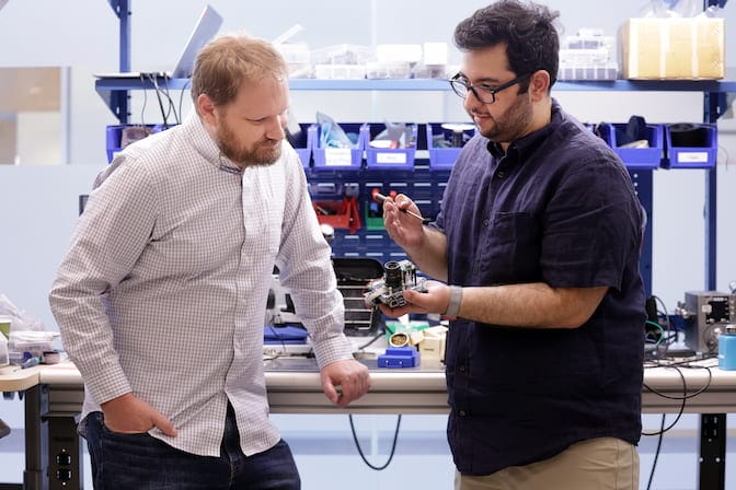 : Two men inspect a handheld camera in front of a messy work bench. One man holds a screwdriver and the camera.