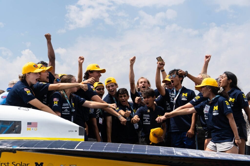 College students cheer while standing behind a vehicle they built.