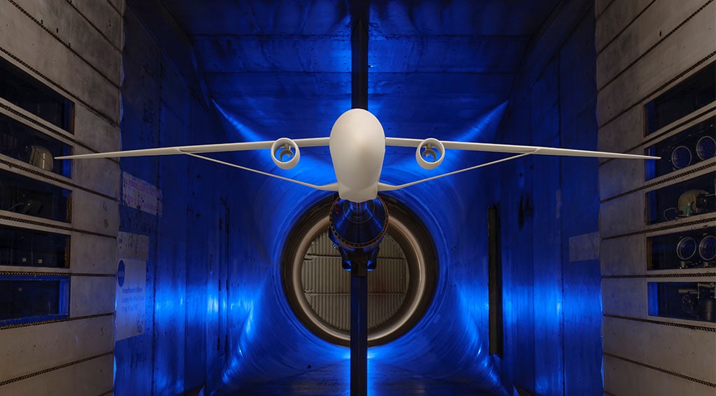 Graphic rendering displaying the front view of an aircraft model with extended wings in a blue-lit wind tunnel.

