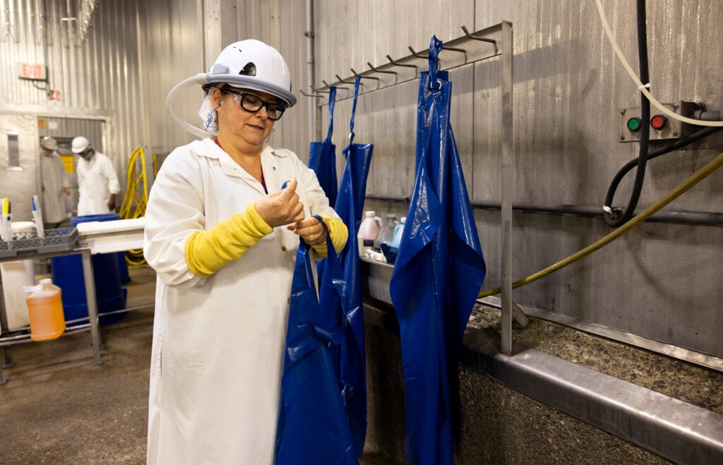 Chaidez wears a white coat and her air curtain headgear, resembling a hard hat, as she prepares to don a blue apron.