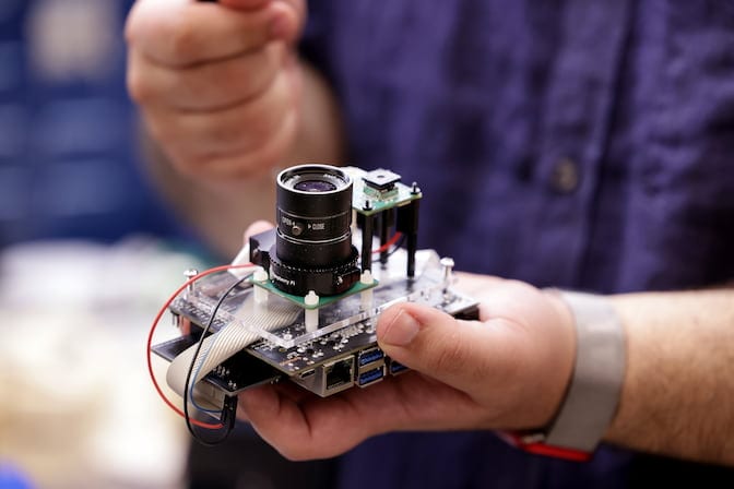 Someone holds a palm-sized camera, which has a long cylindrical lens and a square lens on an elevated platform. The cover of the camera has been removed, revealing the internal electrical components.