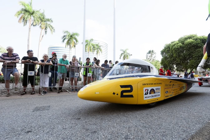 A car, shaped like a yellow bullet with a white cockpit and solar panel array on top, is parked on a paved road in an urban area. Spectators stand behind a nearby fence to watch the race's start. 