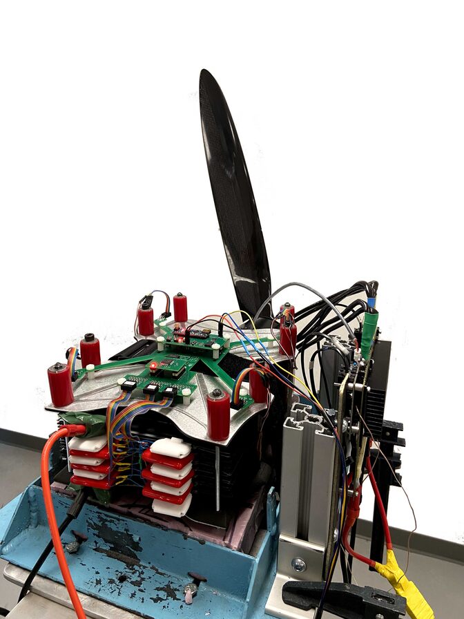 A battery sits on a wooden structure, hooked up to a propeller via exposed wires and electronic boards.