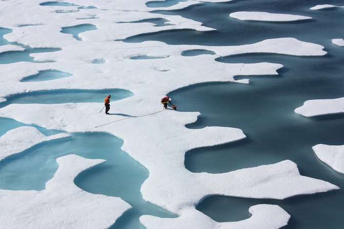 Two scientists walk between blue puddles of slush that speckle the bright white ice sheet. From the aerial vantage point, multiple puddles make the ice resemble holey cheese.