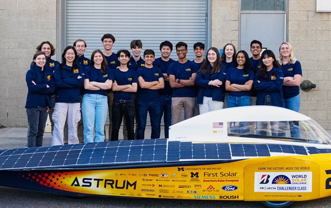 Members of the race crew for the American Solar Challenge stand behind Astrum, which looks like a yellow, bullet-shaped car. The car's top is lined with blue solar panels.