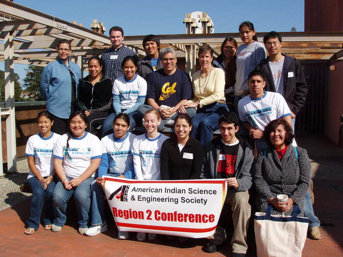 A group of 18 students and instructors gathers in bright sunlight on a brick patio.