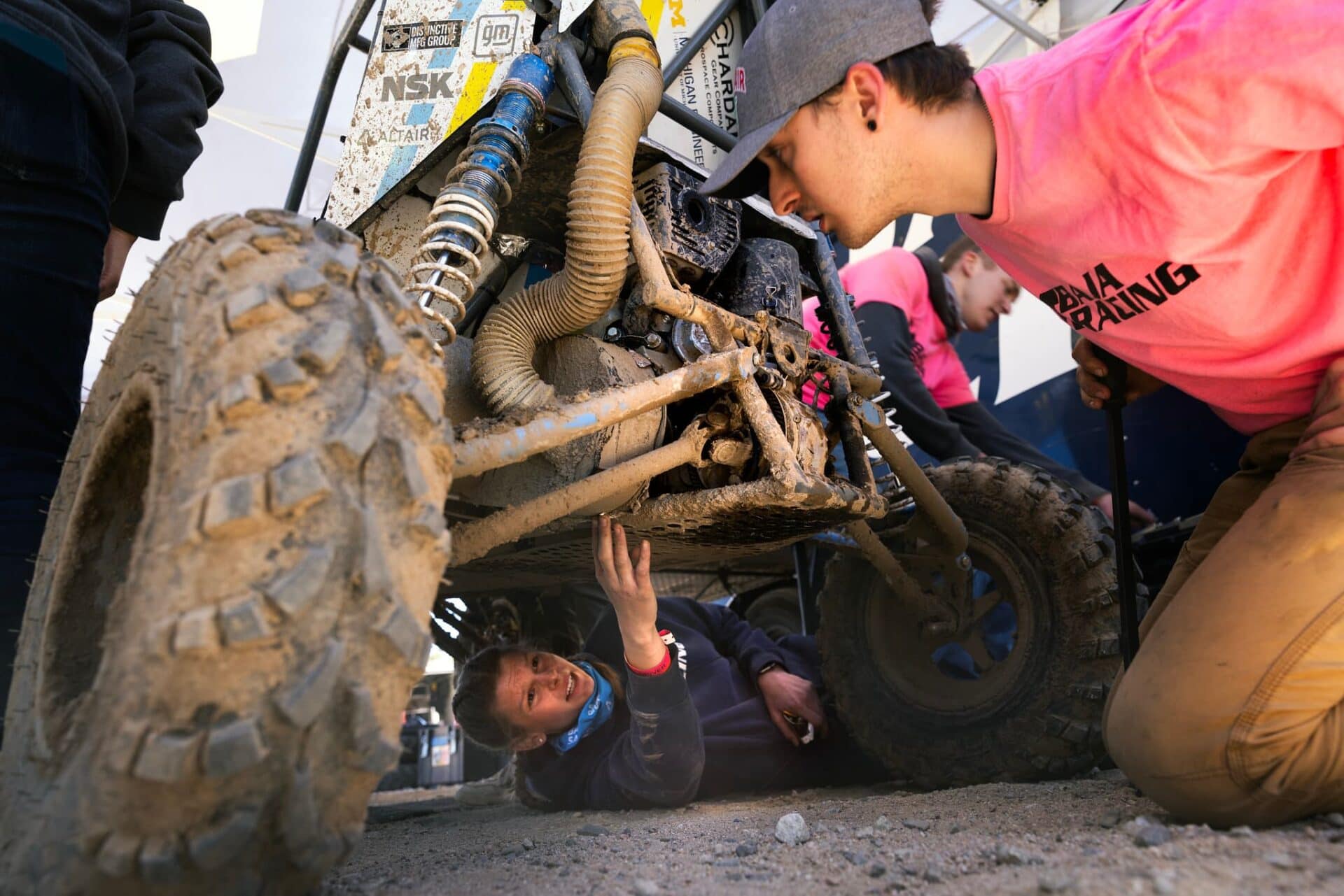 A student crawls underneath the team's dust-caked car, coating herself in dust in the process. Another student kneels down next to the car to get a better view of the engine.