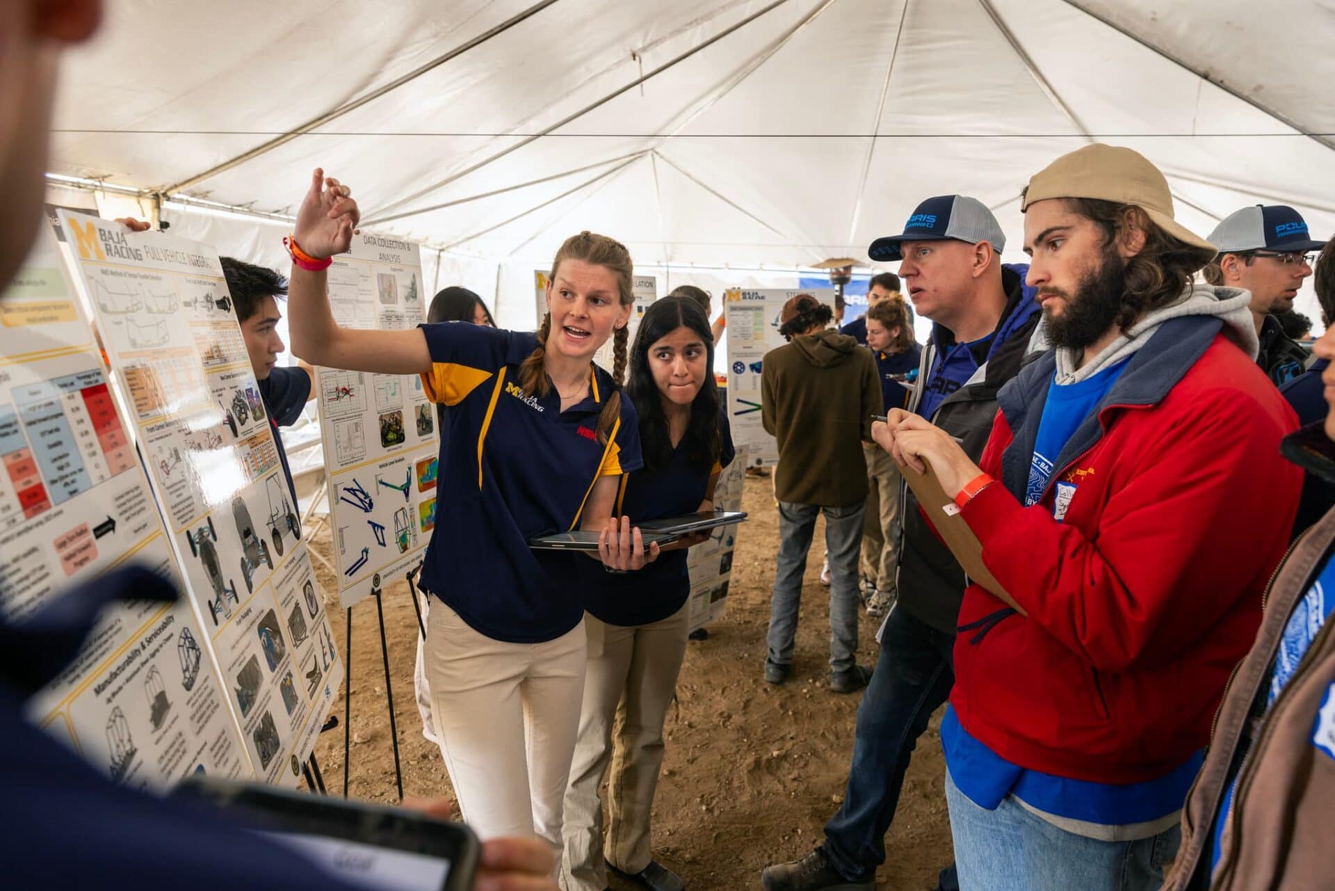 Standing in a large white tent, Lindblom gestures toward a poster on an easel as judges listen with clipboards in hand. Her teammate stands nearby, nervously eyeing the graphs and diagrams on the poster.