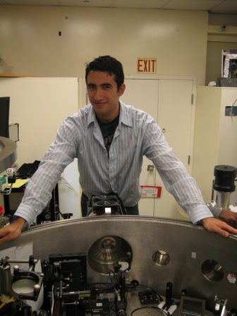  A young man leans over a curved stainless steel enclosure, with lenses and other equipment inside.