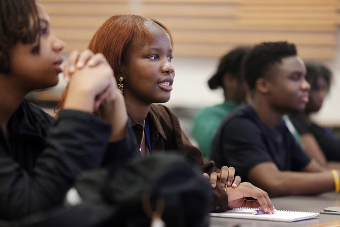 Students attentively listening in a classroom, focused on Aisha Jagne in the foreground.