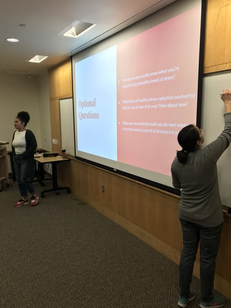 A young woman in red sneakers speaks in front of a projection screen. The current slide asks: "How do you personally know you are experiencing unhealthy levels of stress?" "What healthy forms of stress reduction have worked for you in your first year? How about now?" and "What one new method could you do next week to try to feel more in control of stressors in your life?"