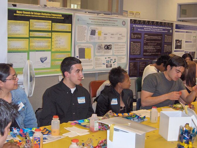 Students share a meal around a long table with academic posters in the background. 