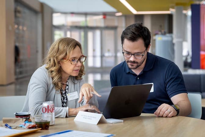 Two workshop attendees sit at a table discussing and working on a laptop together.