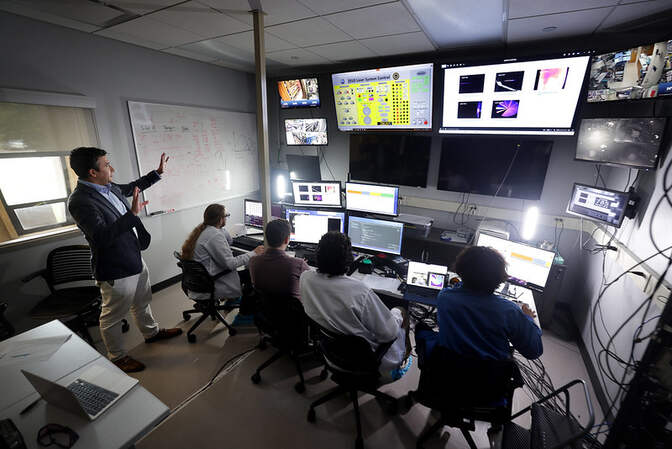 Screens glow in a darkened room. Dollar is speaking and gesturing at the information, while the four student researchers are seated at a table, facing the monitors.