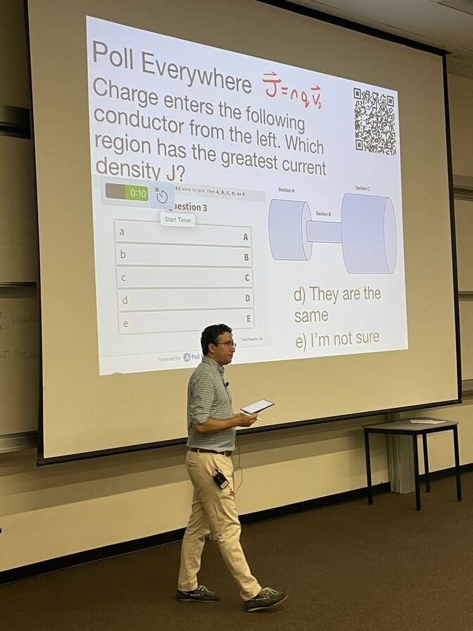 The professor is wired up with a tablet computer and a lavalier mic in front of a projection screen. The quiz question asks students to identify where the current density will be highest in an image that resembles an asymmetric dumbbell. Students have the option of choosing "I'm not sure."