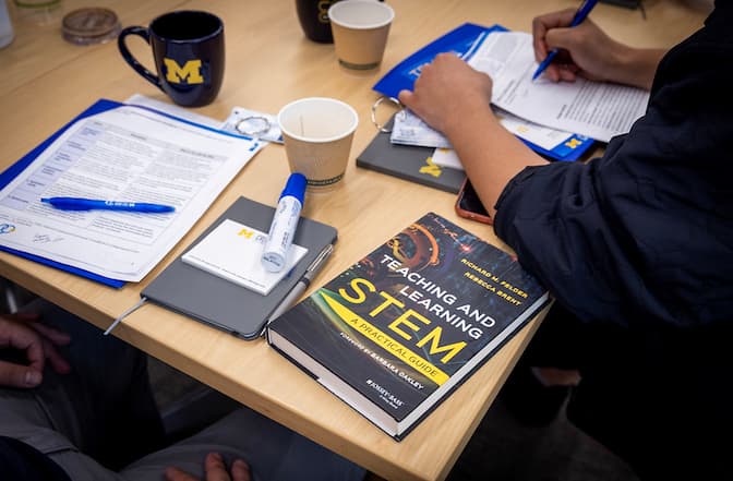 Items on a table including a blue folder with papers, a book titled "Teaching and Learning STEM," notebooks, pens, coffee cups, and mugs with a maize block "M." A person is writing on papers.