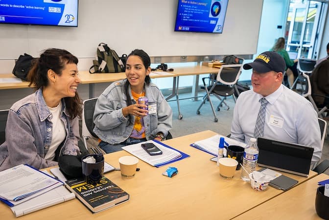 Three individuals at a table during a workshop, engaged in conversation with notebooks, coffee cups, and electronic devices on the table.