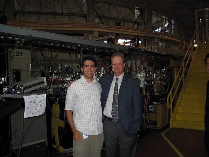 An older man in a suit has an arm around a smiling young man, with metal equipment gleaming behind them and a yellow staircase off to the right.