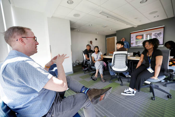 Professor Todd Allen in a blue shirt speaks to a group of high school students in a modern meeting room.