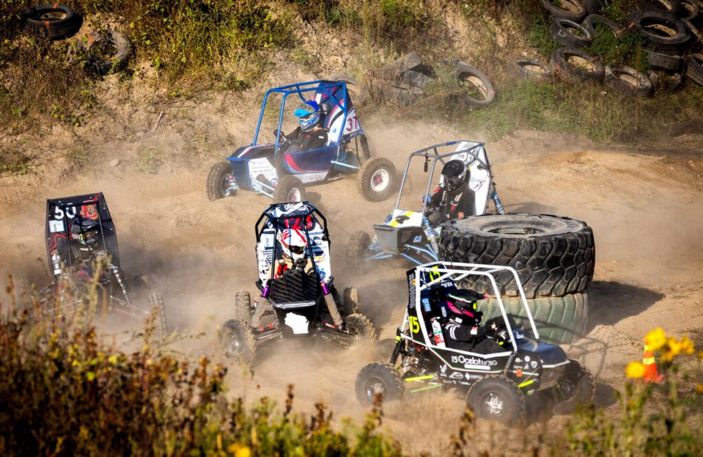 Five baja cars spray dust into the air as they speed around a stack of two tractor tires. The white Michigan car cuts past the blue and pink car from John Hopkins and Cornell's black car. Two other black cars with colored decal maintain their lead.