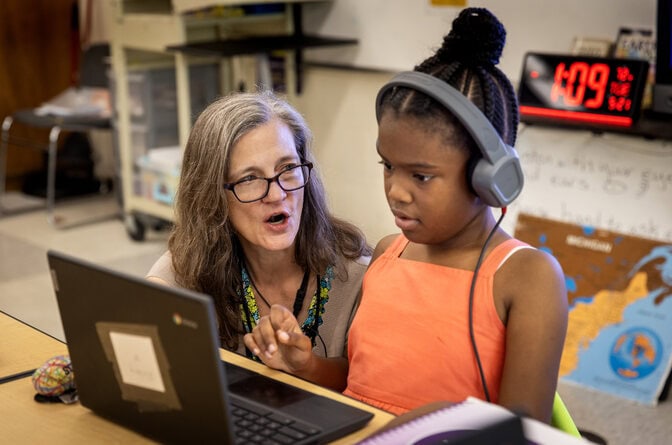 A woman kneels next to a young student sitting at her desk. The woman looks at the student, lips rounded in a vowel sound. The student looks at the screen of the laptop on her desk.