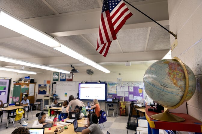 The American flag hangs above a small globe in a corner of Bruzzano's classroom. Below the flag and globe, students work on their laptops as Bruzzano leans over the shoulder of a student toward the back of the room.