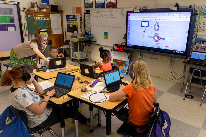 Four students sitting at a cluster of desks work on their laptops. One student has turned toward a large smartboard hanging on the classroom wall behind her. The smartboard shows the shape and direction of the magnetic field. Nearby, the teacher leans over a student's shoulder to point out something on the student's computer screen.