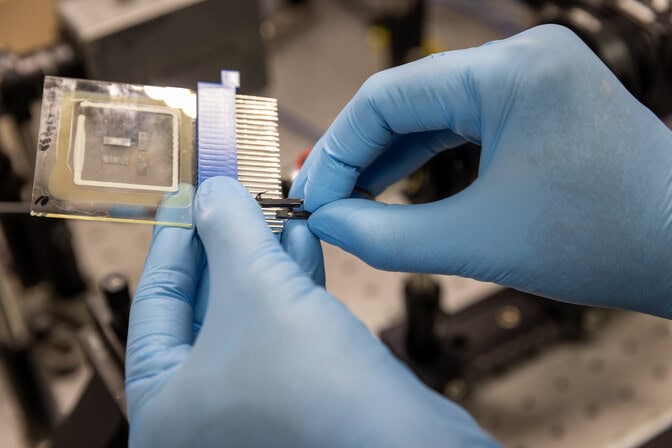 Gloved hands connect a wire to the OLED, two sheets of glass about ten by ten centimeters, with four, one centimeter long translucent gray strips sandwiched between. The silver barrel of a microscope points at the OLED. Housing insulation is out of focus behind.