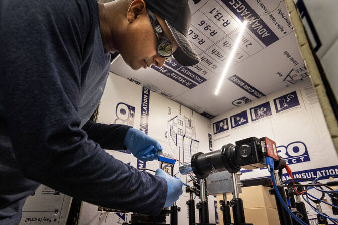 : A researcher wearing safety glasses, gloves and a baseball cap connects a wire to the OLED, two sheets of glass about ten by ten centimeters with translucent gray strips sandwiched between. A mounted, horizontally positioned microscope points at the OLED. Housing insulation surrounds the experimentation area.