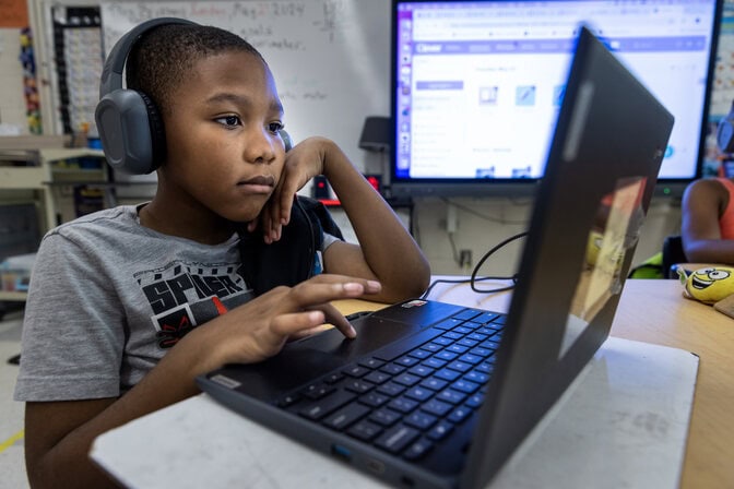 A young student wearing headphones concentrates on the digital coursework displayed on the screen of their laptop.