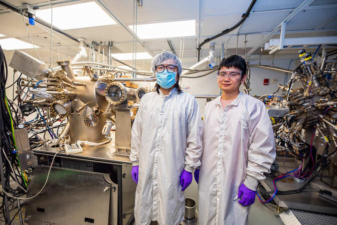 Pan and Bingxing stand in a lab. Two devices in the background are full of portholes, pipes and wires.