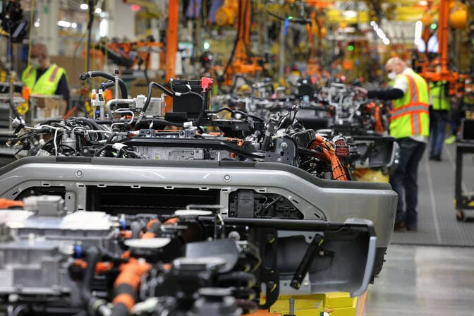 A long line of pickup chassis are shown on the assembly line, with workers in goggles and safety vests working nearby.