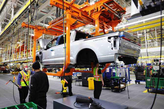 Two autoworkers stand beside the frame of an elevated all-electric pickup truck body on the assembly line, while another works beneath it.