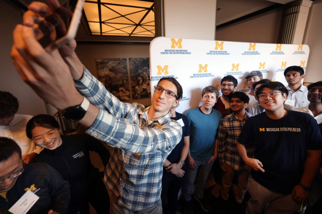 Students gather around Sam Altman for a group "selfie" photograph. Behind them is a University of Michigan backdrop.