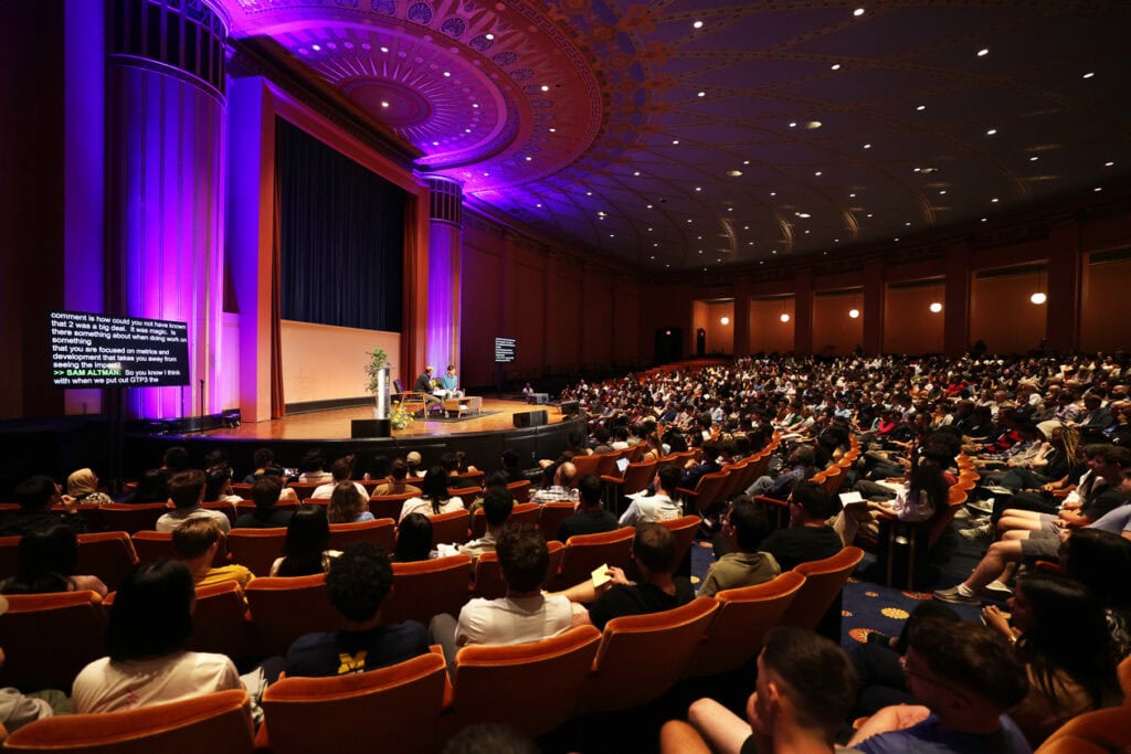 A wide photograph showing a packed theatre. In the center of the photo, Sam Altman and Dan Feder are seated on stage.