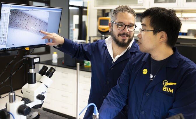 Two men in blue lab coats stand in front of a microscope. One of the men is pointing at a computer screen that displays the magnified squid skin, which looks like a pale tube speckled with dark red dots.