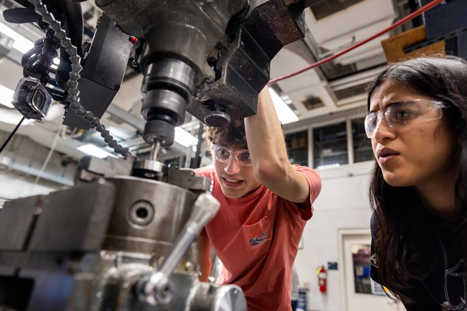 Two students wear safety gear and operate a machine mill