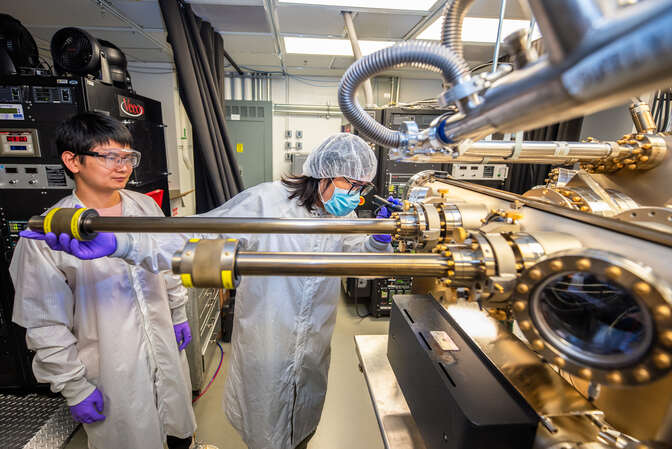 Two scientists in white lab coats and purple gloves at a machine with many portholes, some equipped with long metal tubes.