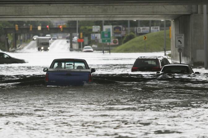 A pickup truck, submerged in water halfway up the driver’s door, is seen from the back attempting to drive beneath an overpass, while nearby cars appear stuck in the flooding.