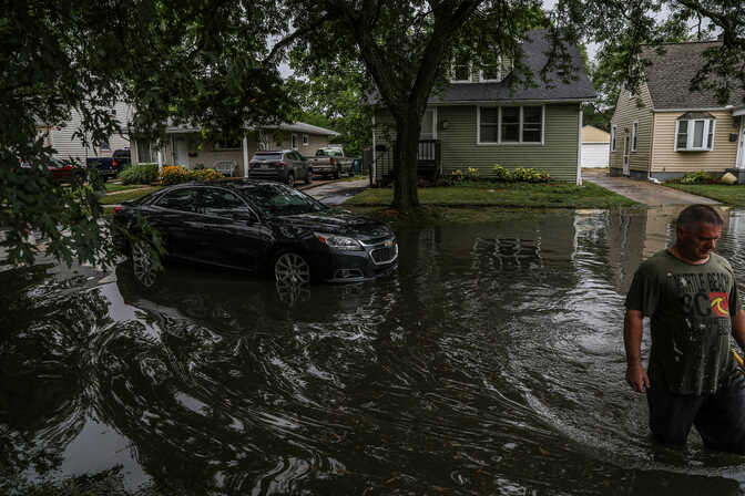 A man in a t-shirt wades across a residential street in flood waters above his knees, carrying a yellow tool. A stalled car is behind him in the middle of the street.